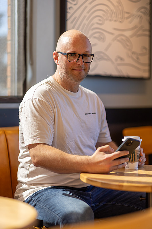 Paul sitzt mit Handy und Kaffee an einem Tisch in einem Café